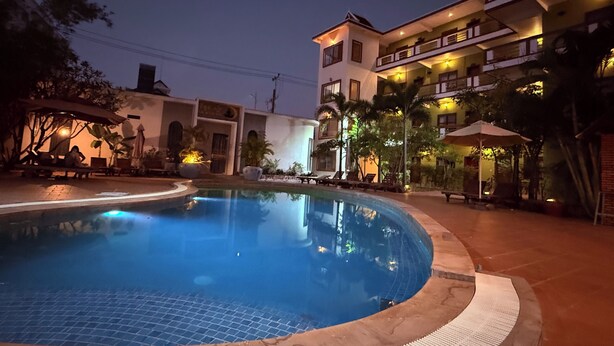 Una piscina rodeada de sillones de descanso y sombrillas, con un edificio de balcones e iluminación exterior al fondo.