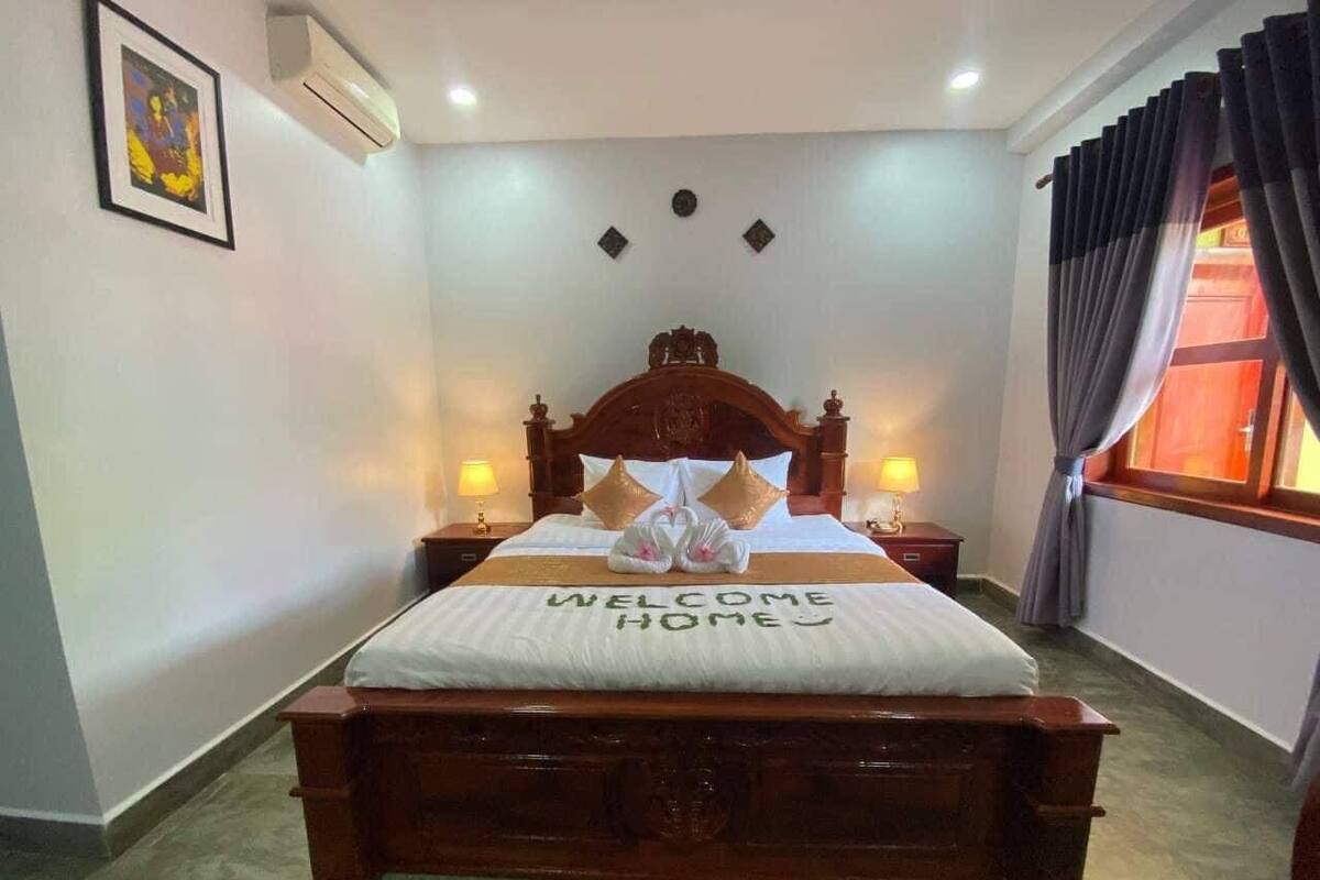 Een net opgemaakt bed met een “WELCOME HOME” bord, een houten hoofdbord, twee bedlampen en een raam met gordijnen.