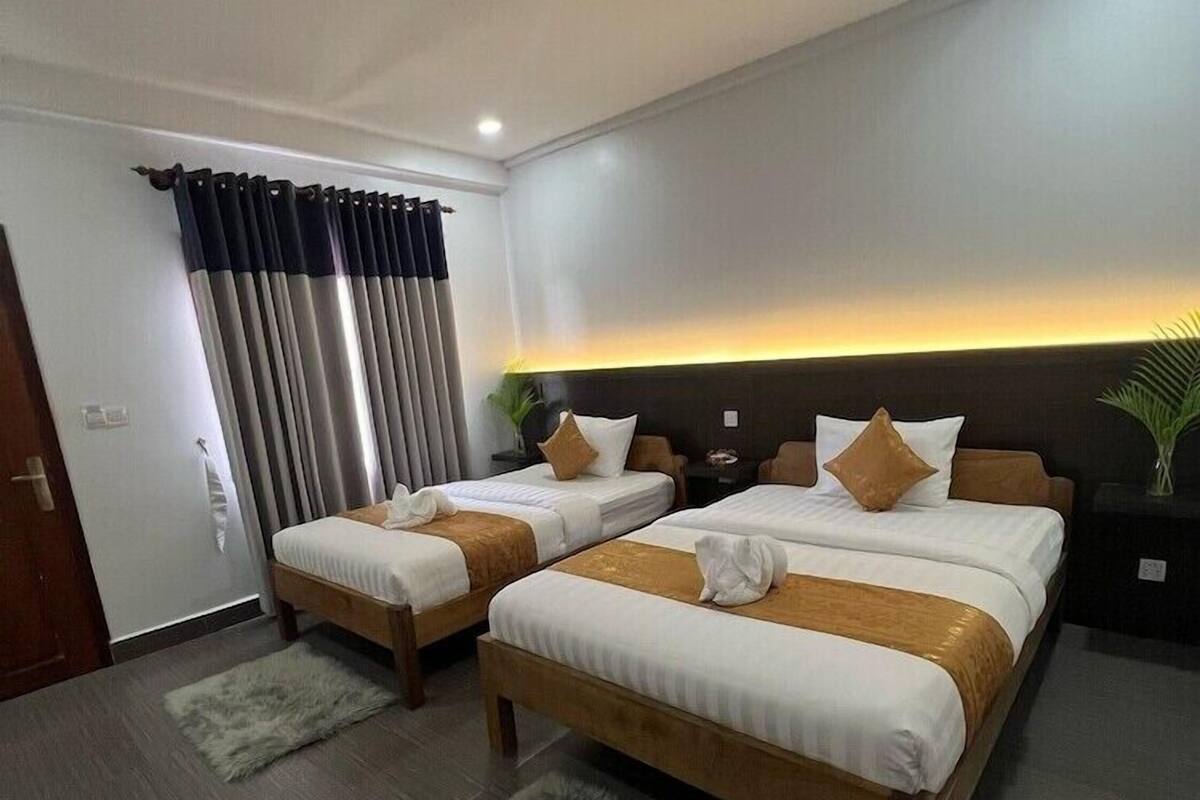 Een hotelkamer met twee bedden, een groot raam met gordijnen, een nachtkastje en een plant.