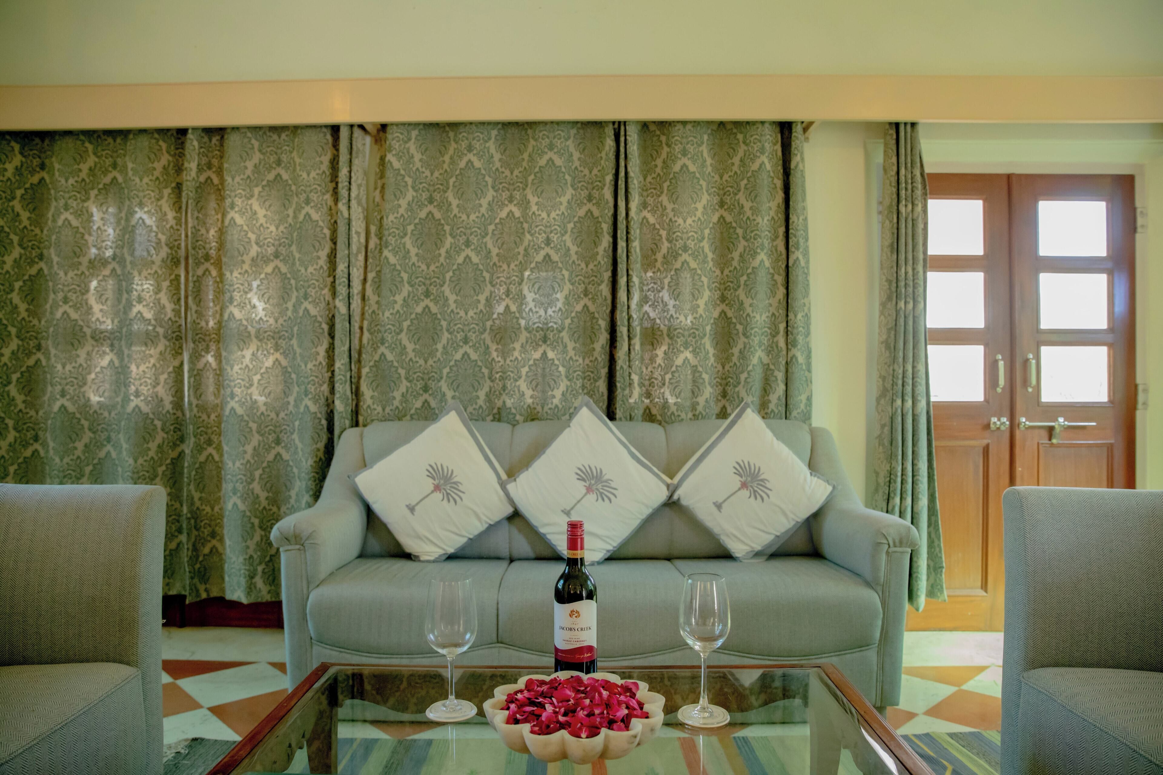 A living room with a sofa, two armchairs, a glass coffee table with a wine bottle and glasses, and a patterned curtain backdrop.