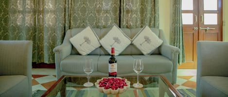 A living room with a sofa, two armchairs, a glass coffee table with a wine bottle and glasses, and a patterned curtain backdrop.