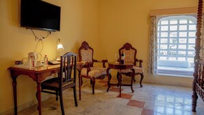 A room with a television mounted on the wall, two wooden chairs with patterned upholstery, a small round table with a wine glass, and a window with a view of trees. - Shahpura Abhaneri Resort (Baswa)