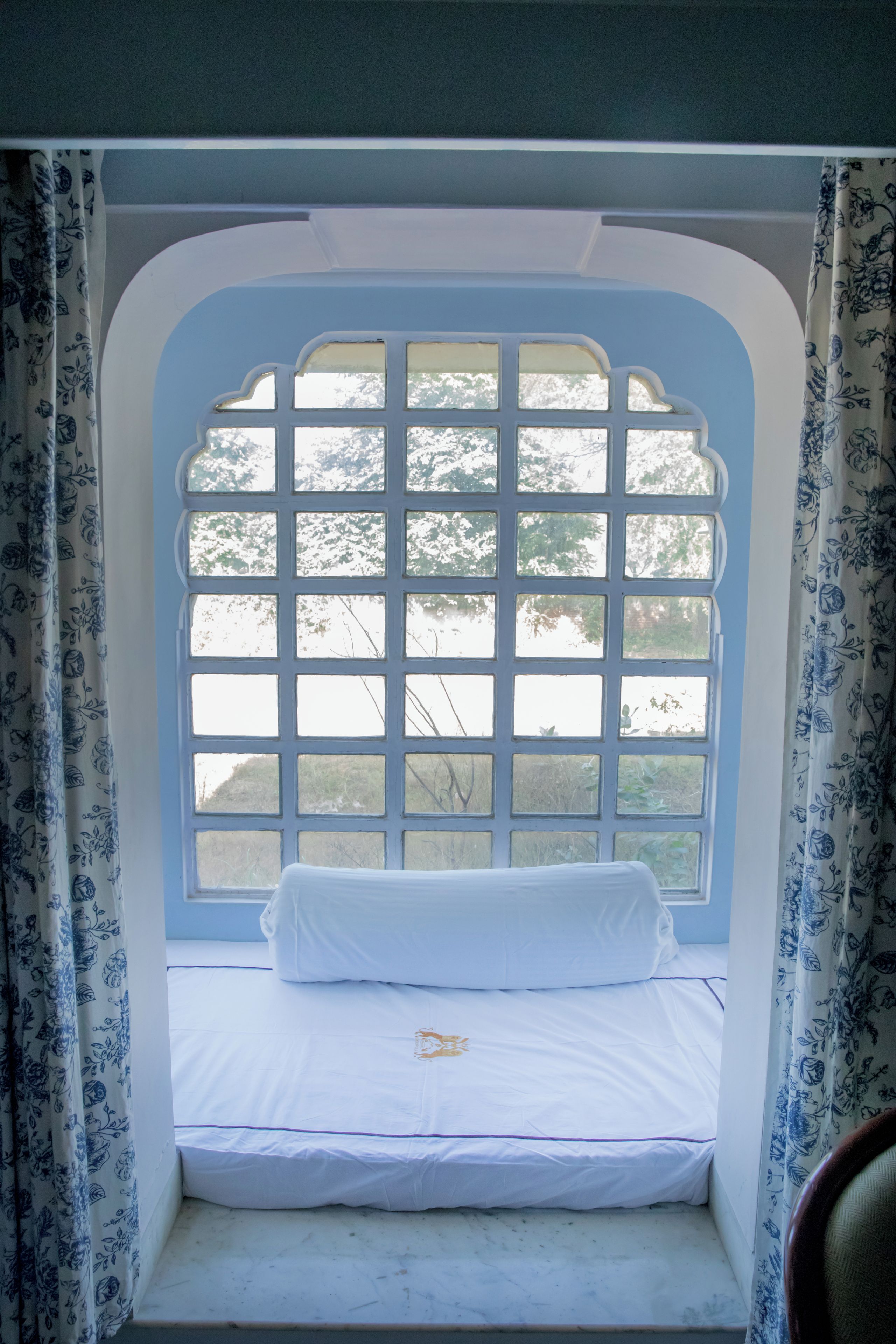 Ein Zimmer mit Fensterbank, ausgestattet mit einer weißen Matratze und Kissen, umgeben von blumigen, blauen Vorhängen.