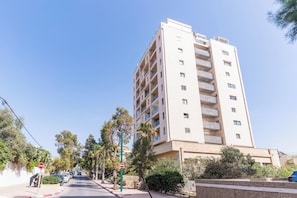 A multi-story residential building with balconies, a clear sky, and a street with parked cars and a pedestrian.