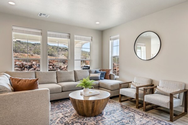 A modern living room with a sectional sofa, two armchairs, a round coffee table, and large windows offering a view of a rocky landscape.