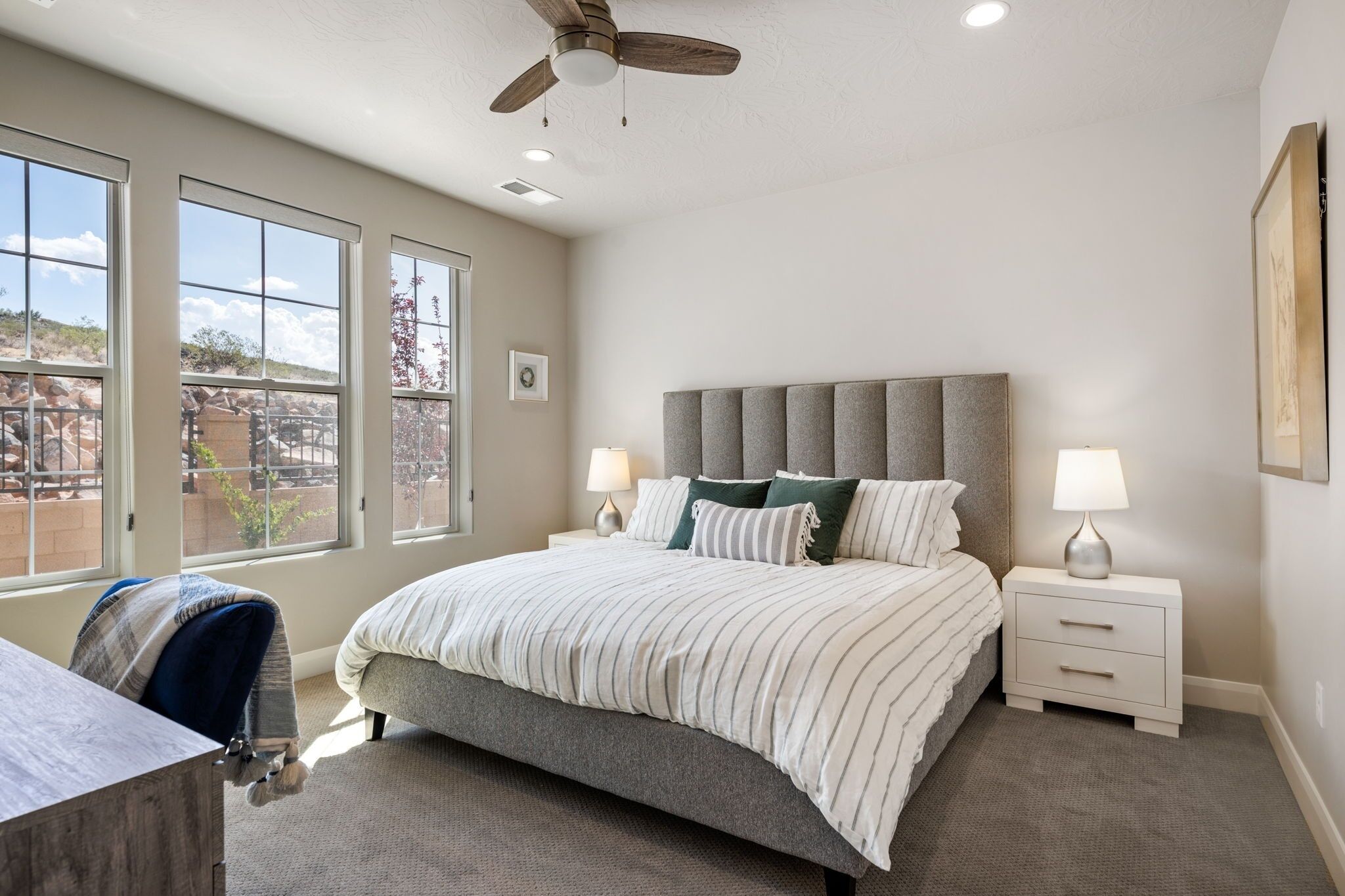 A bedroom with a large bed, two bedside tables with lamps, a ceiling fan, and large windows with a view of the outdoors.