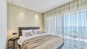 Een moderne slaapkamer met een groot bed, een nachtkastje en uitzicht op zee door dunne gordijnen.