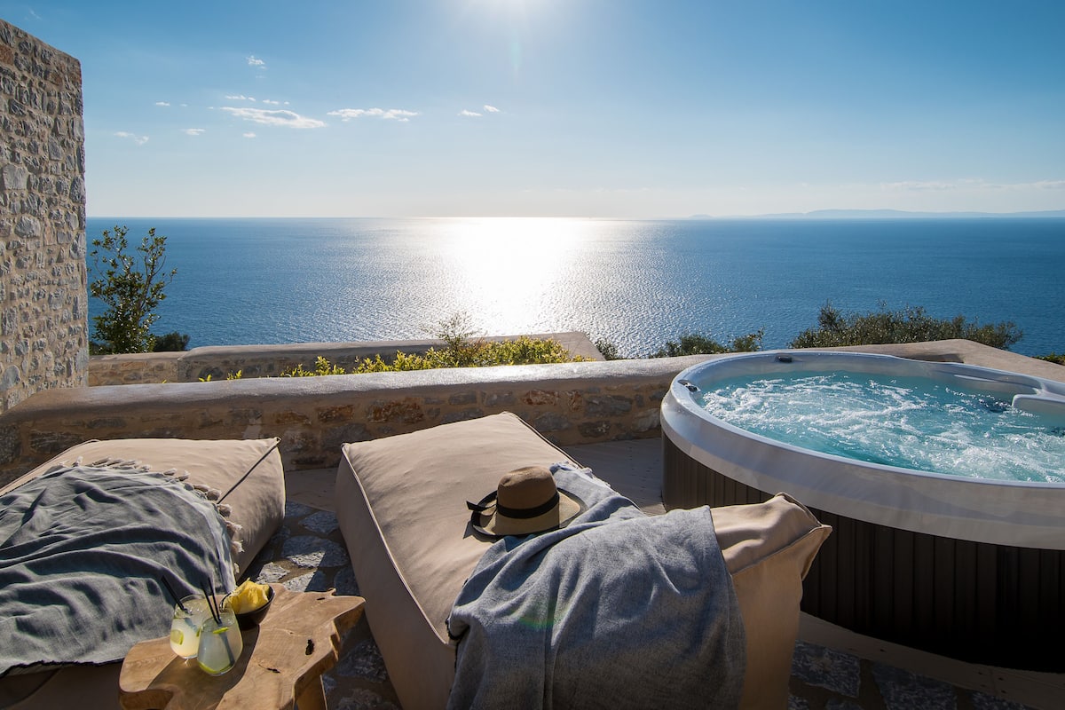 A sunlit outdoor seating area with a view of the sea, a hot tub, and a stone wall.