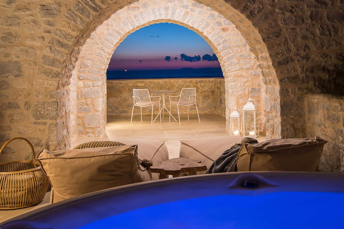 A stone archway leading to a terrace with a view of the sunset over the sea, furnished with a table and chairs, and a hot tub in the foreground.