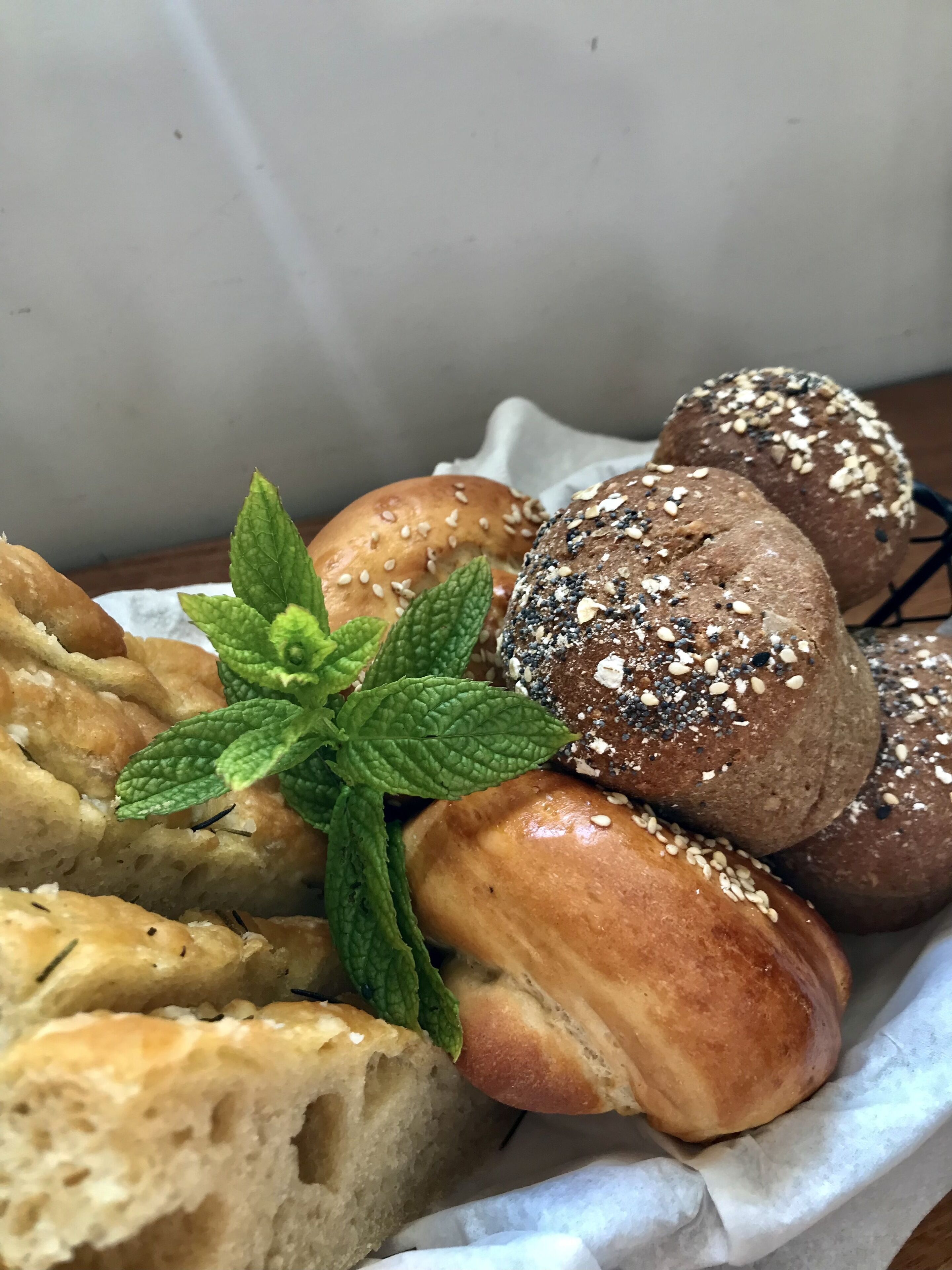 A basket of various breads and rolls, including sesame and poppy seed varieties, with a mint leaf garnish.