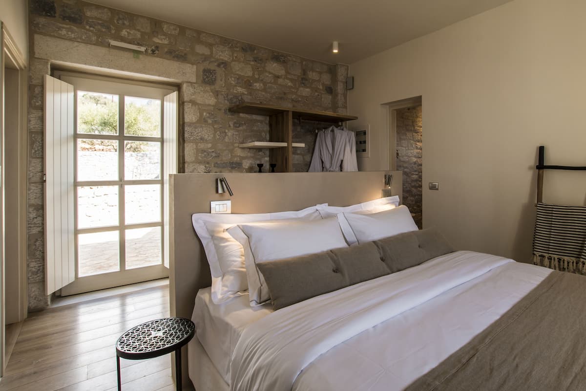 A bedroom with a stone wall, a bed with white bedding, a small table, and a wooden ladder.