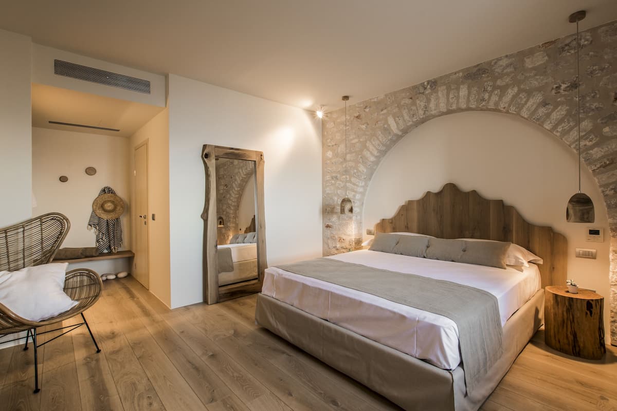 A bedroom with a large bed, a wooden headboard, a stone wall, a wooden floor, a wicker chair, and a bedside table.