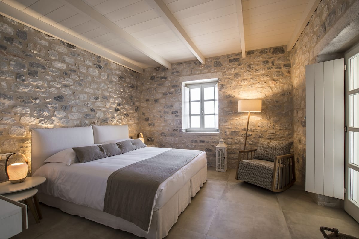 A bedroom with a stone wall, a large bed, a nightstand, a chair, and a window.