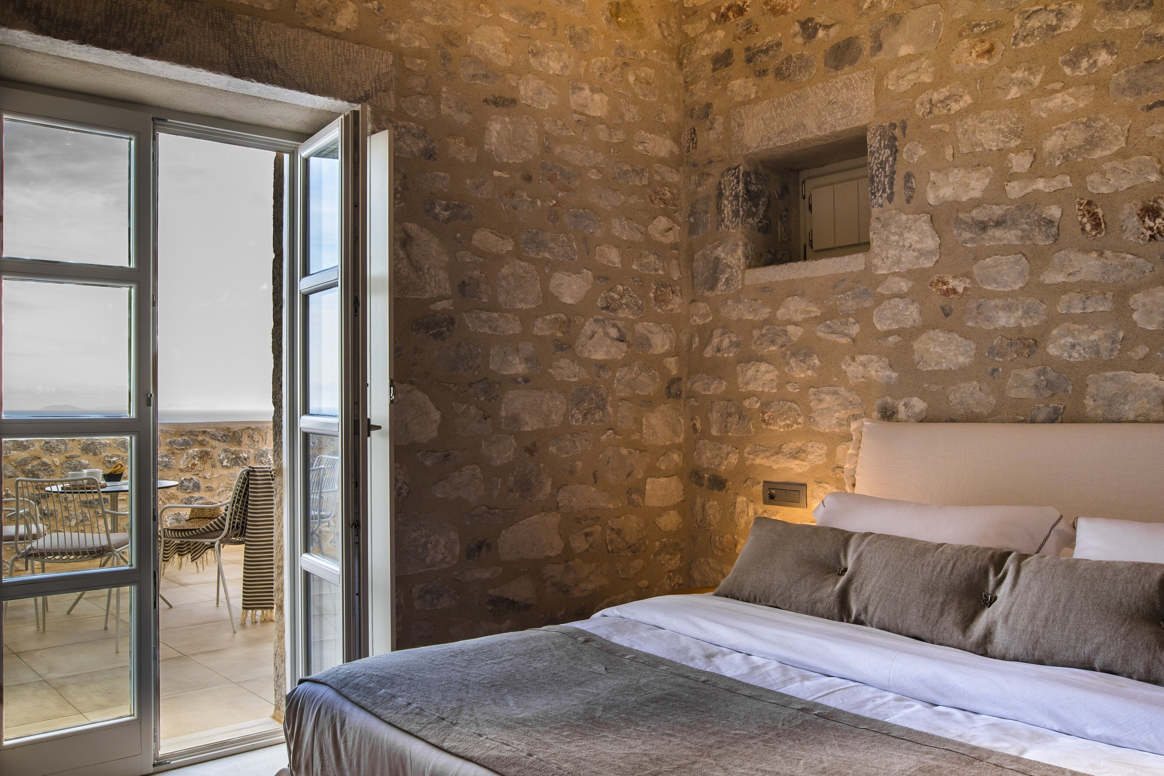Un dormitorio con pared de piedra, una cama con ropa blanca y vistas a un patio exterior con sillas y una mesa.