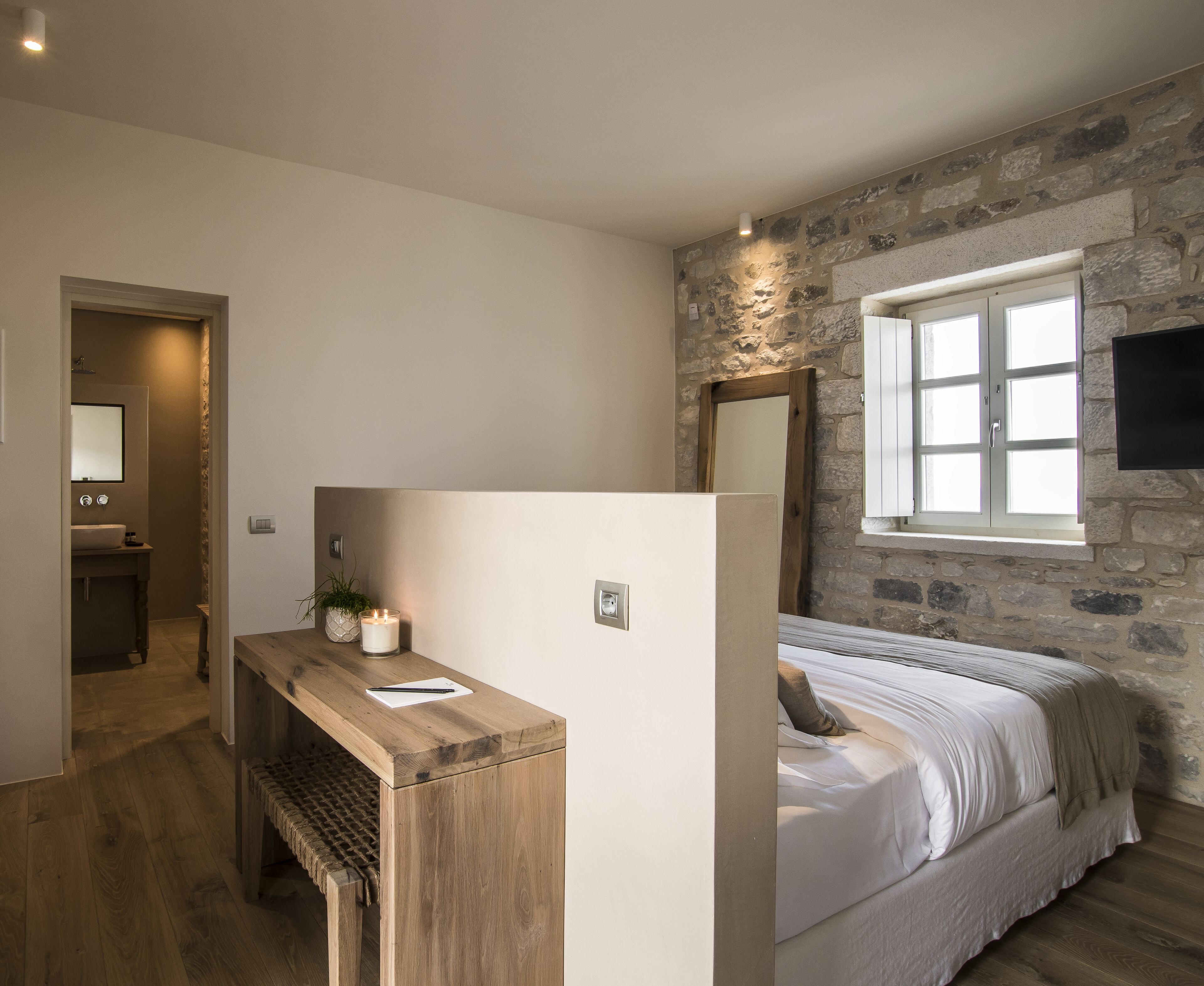 A bedroom with a stone wall, a wooden desk, a bed, and a window.