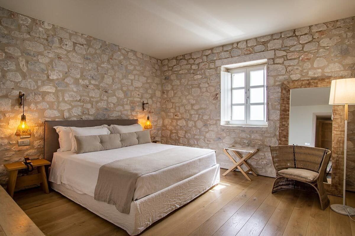 A bedroom with a stone wall, a bed, a chair, and a window.