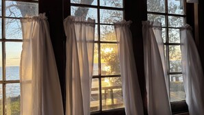 Ein Zimmer mit großen Fenstern, weißen Vorhängen und Blick auf Bäume und ein Gewässer.