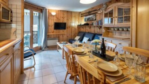A wooden-furnished dining area with a table set for two, a television mounted on the wall, and a balcony with sliding doors. - Husky Cosy 2 Bed Alpine Apartment Views & Balcony (Saint-Gervais-les-Bains)