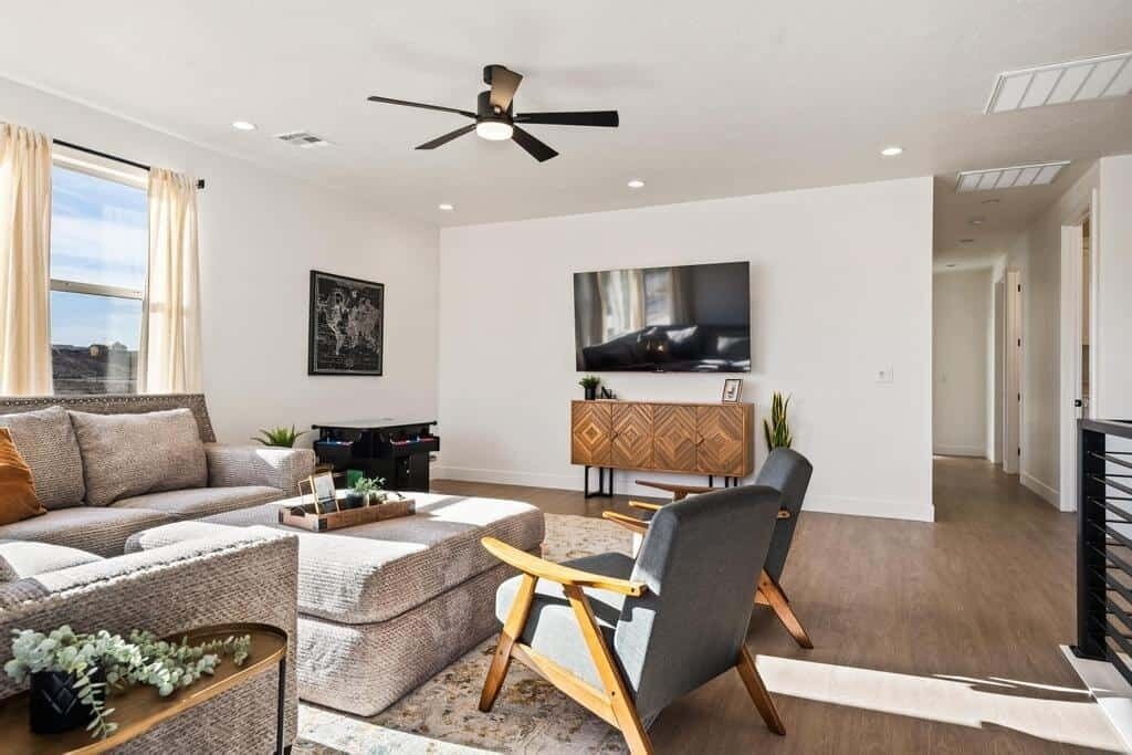 A modern living room with a sofa, ottoman, and a chair. There is a television on the wall and a ceiling fan.