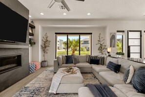 Un salon moderne avec un canapé d’angle, une cheminée et de grandes fenêtres donnant sur l’extérieur.