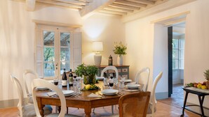 A dining room with a wooden table set for four, surrounded by white chairs. There are wine bottles, glasses, and a fruit bowl on the table. - Anticiana 8 (Lucca)