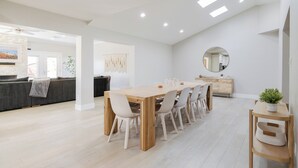 Een ruime eethoek met een lange houten tafel en witte stoelen, een ronde spiegel aan de muur, en een moderne keuken op de achtergrond.