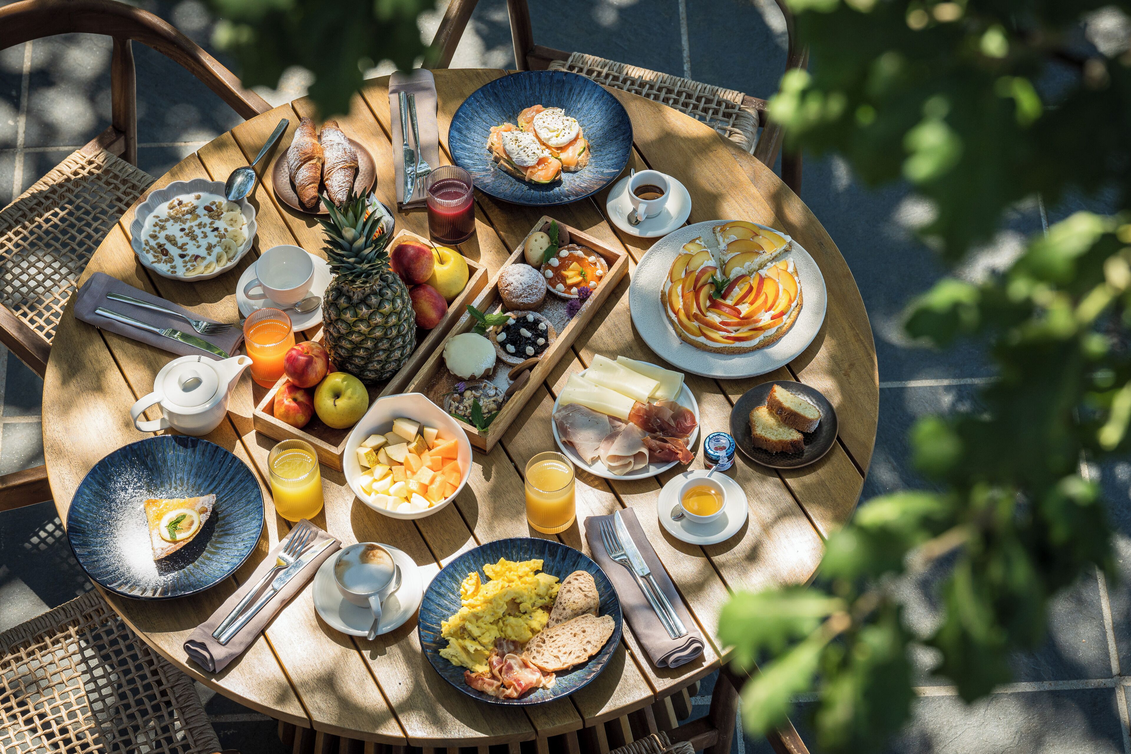 Een ronde houten tafel met ontbijtartikelen, zoals fruit, gebak en drankjes.
