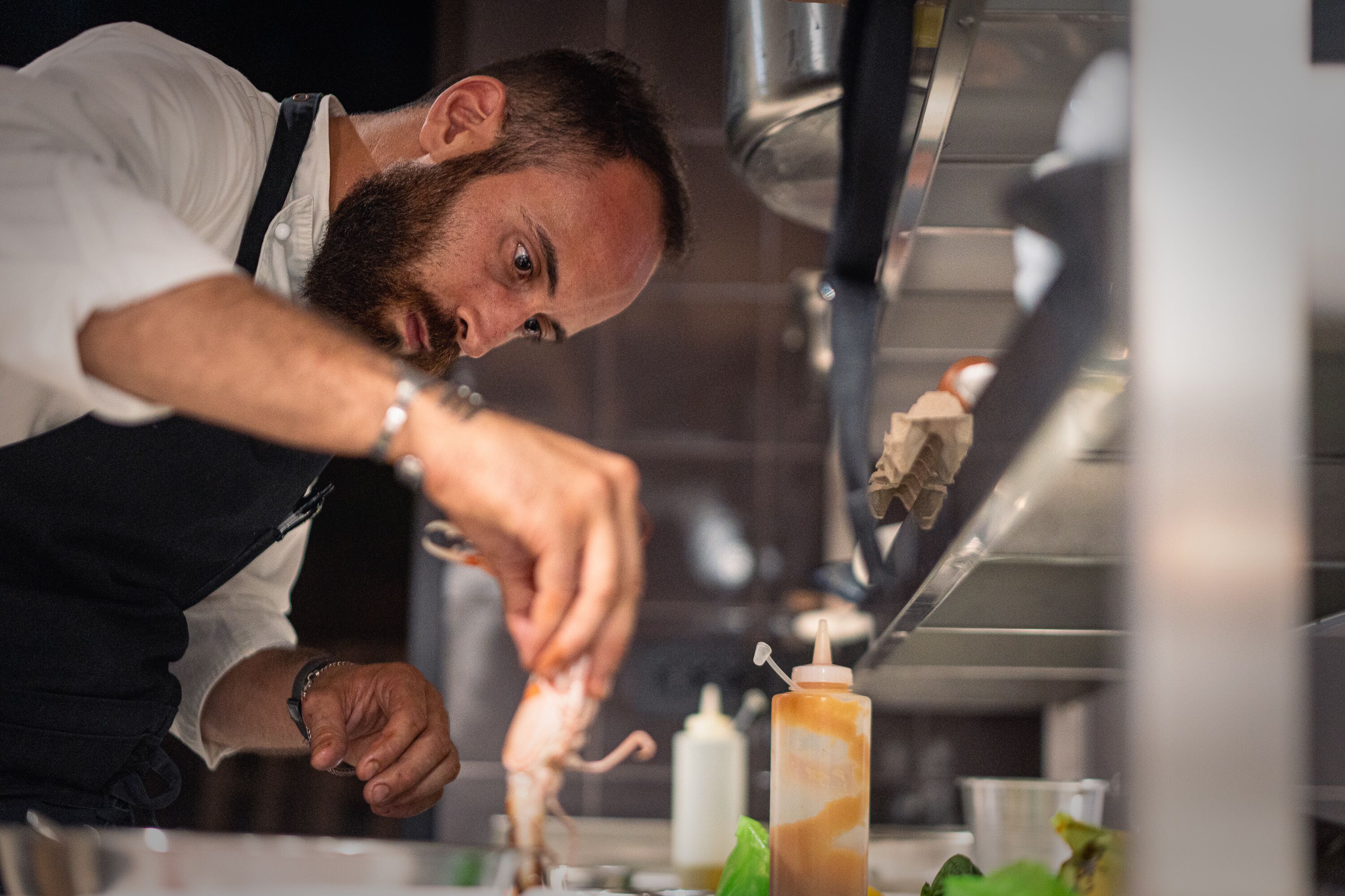 Un chef préparant des plats dans une cuisine équipée d’appareils en acier inoxydable, avec une bouteille de sauce à portée de main.