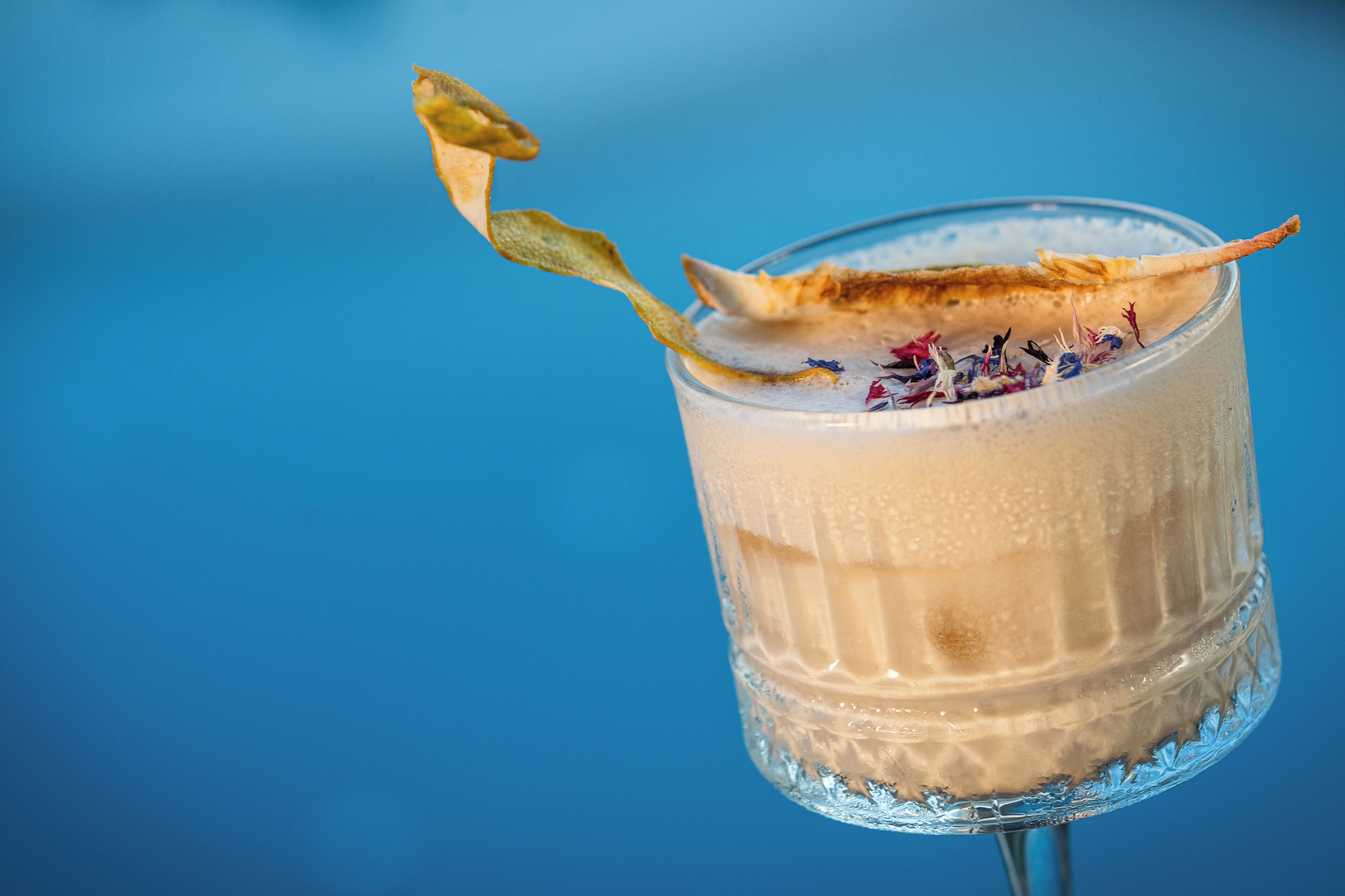 A clear glass with a creamy beverage, garnished with a leaf and flower petals, against a blue background.