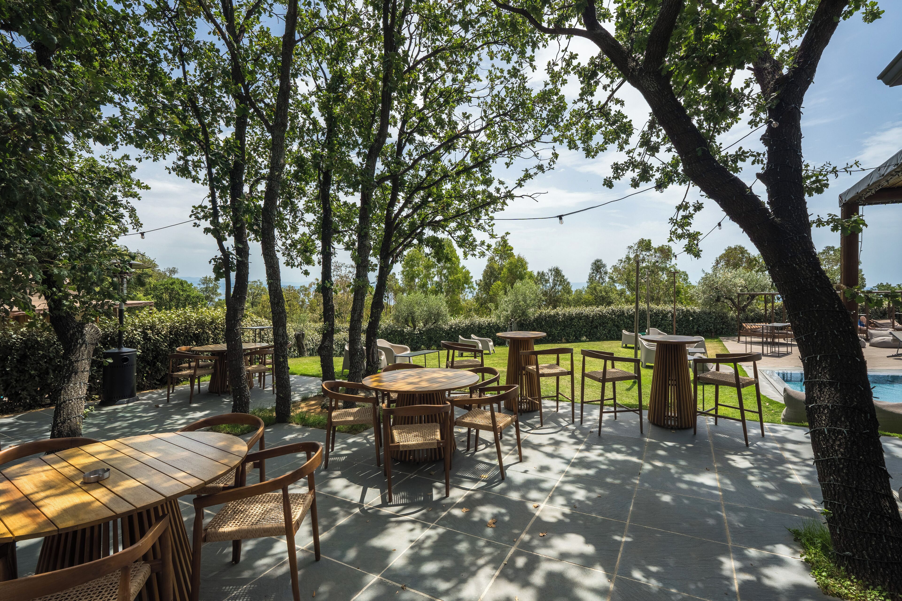 Un'area patio all'aperto con diversi tavoli e sedie in legno, circondata da alberi e una piscina.