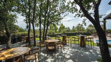 Un'area patio all'aperto con diversi tavoli e sedie in legno, circondata da alberi e una piscina.