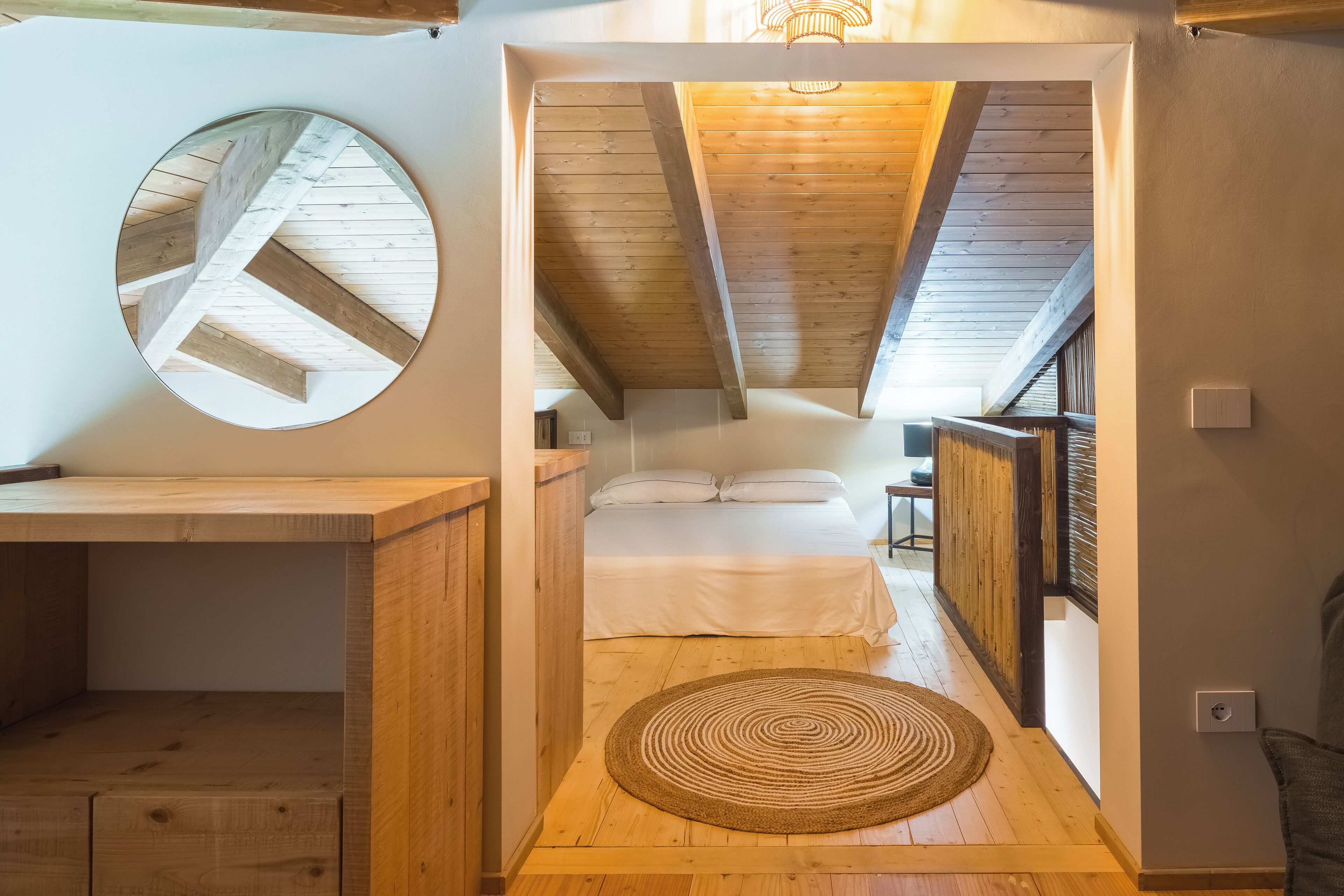 A cozy attic bedroom with a wooden ceiling, a circular mirror, a bed with white linens, and a woven rug on the floor.