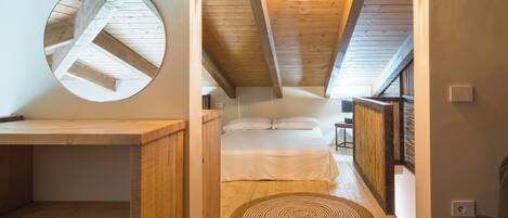 A cozy attic bedroom with a wooden ceiling, a circular mirror, a bed with white linens, and a woven rug on the floor.
