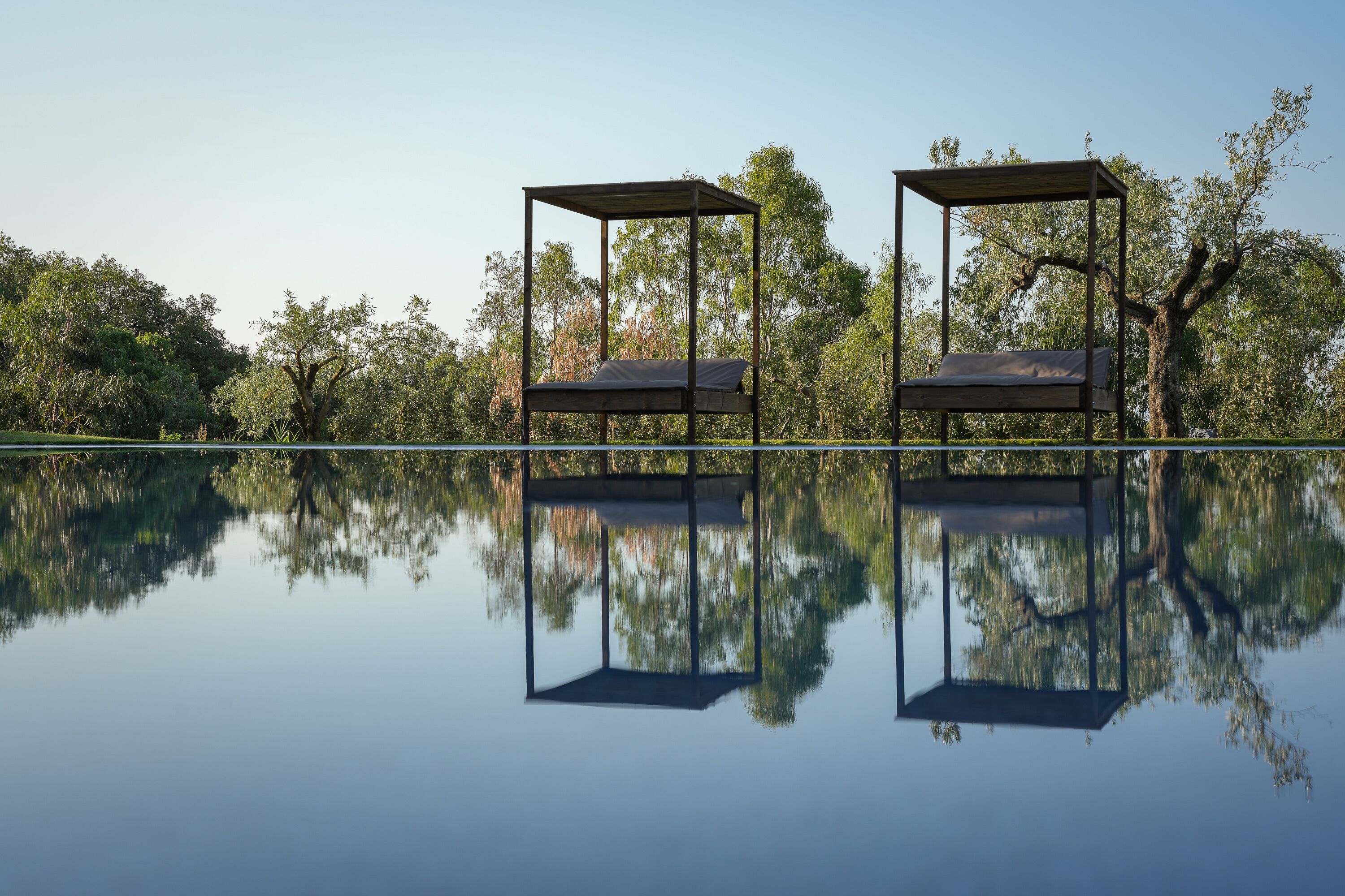 Two outdoor beds with metal frames and wooden roofs, set against a backdrop of trees and a calm body of water.
