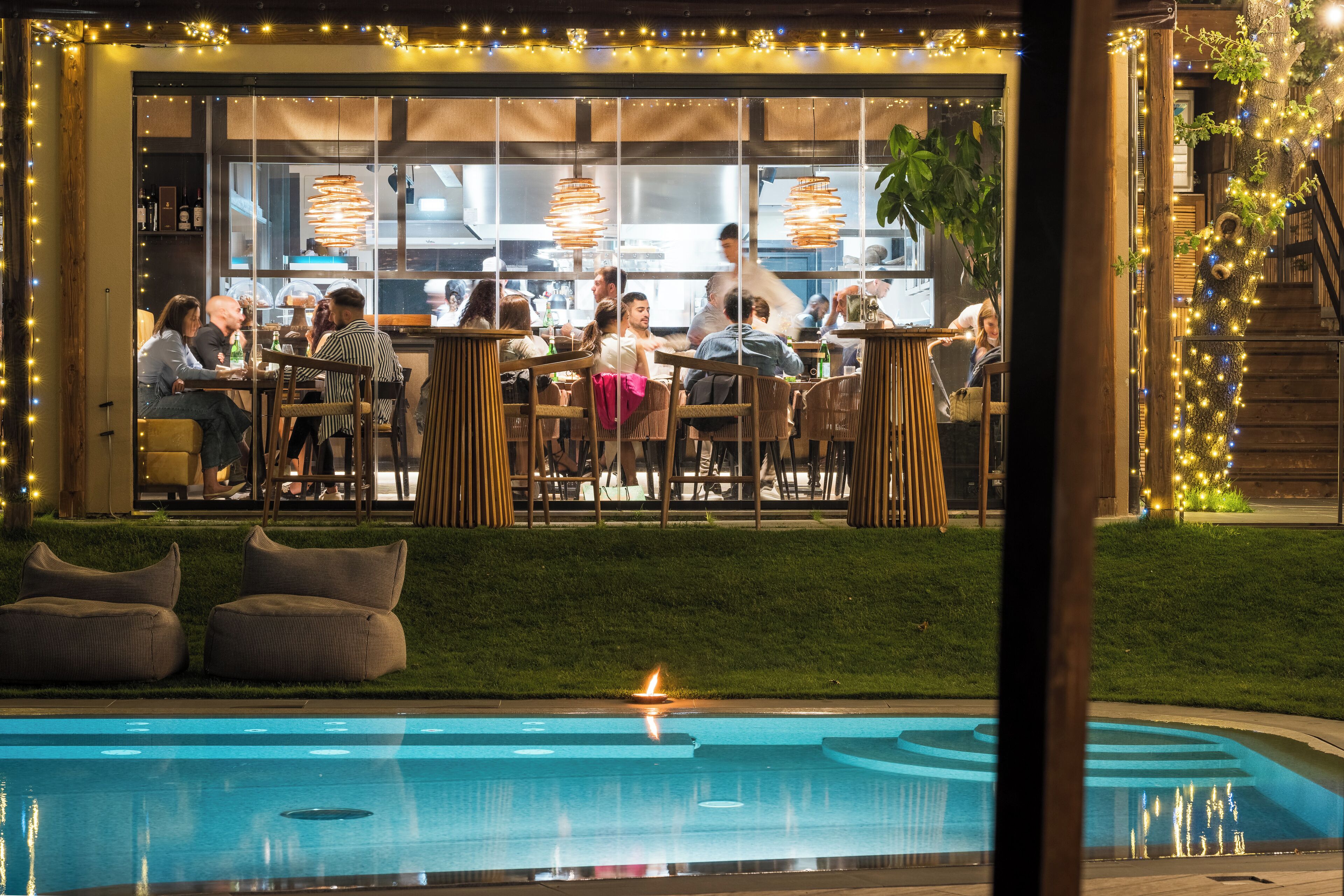 Un espace extérieur aménagé pour les repas, avec des tables et des chaises, une piscine illuminée par une bougie, et une vue sur le restaurant à travers de grandes fenêtres.