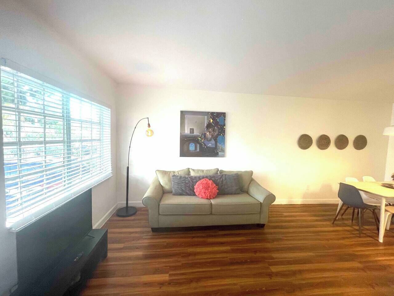 A living room with a sofa, a dining table with chairs, a TV, a floor lamp, and a window with blinds.