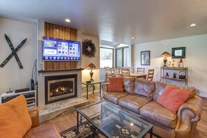 A living room with a fireplace, leather sofa, glass coffee table, and a flat-screen TV mounted above the fireplace.