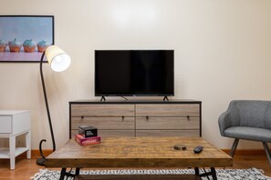 A living room with a flat-screen TV on a wooden stand, a lamp, a coffee table with books and a remote, a grey armchair, and a white side table.