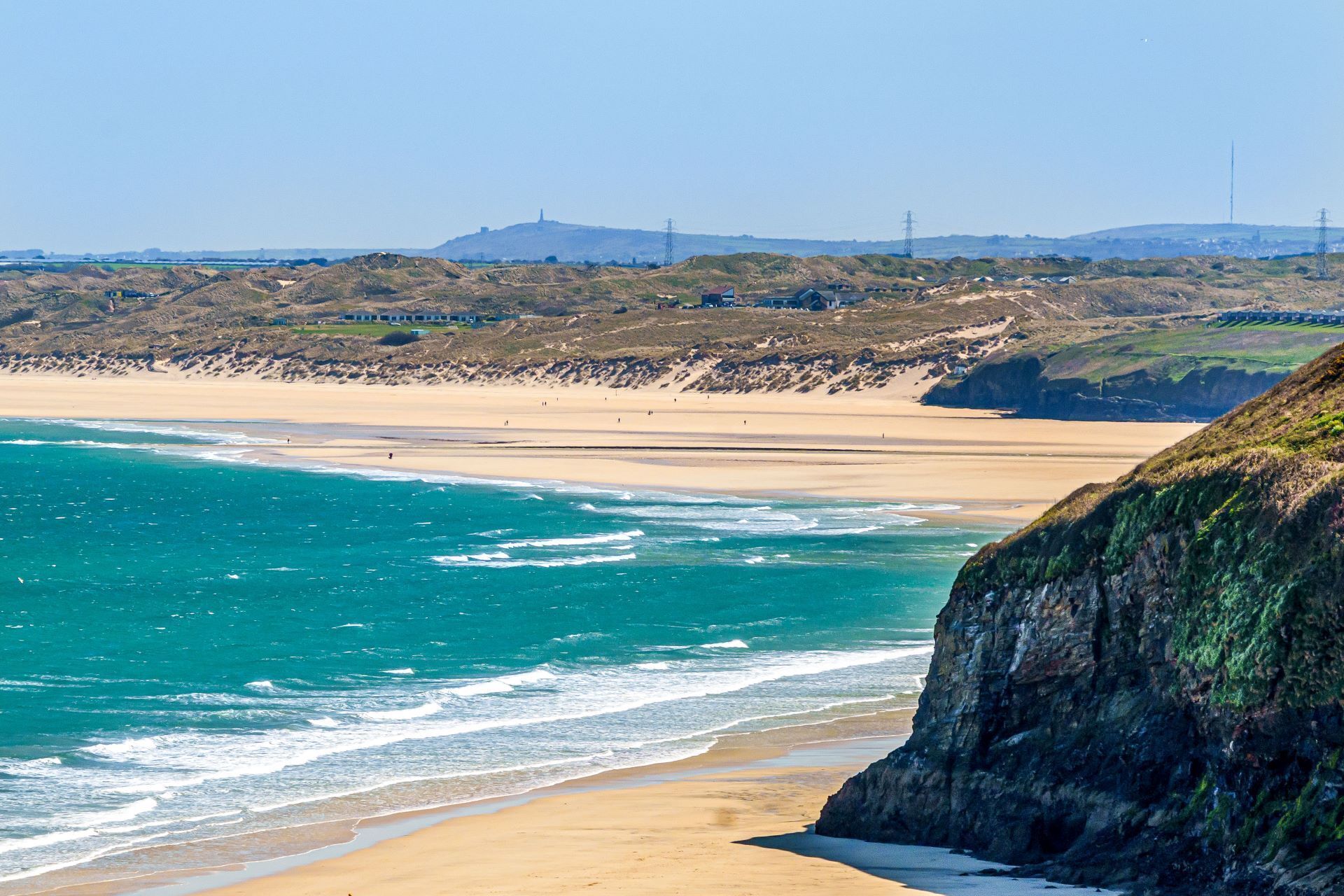 A coastal beach with clear turquoise waters, sandy shores, and a rocky cliff.