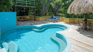 Una piscina con cubierta de madera, sillas de descanso y sombrilla de paja.