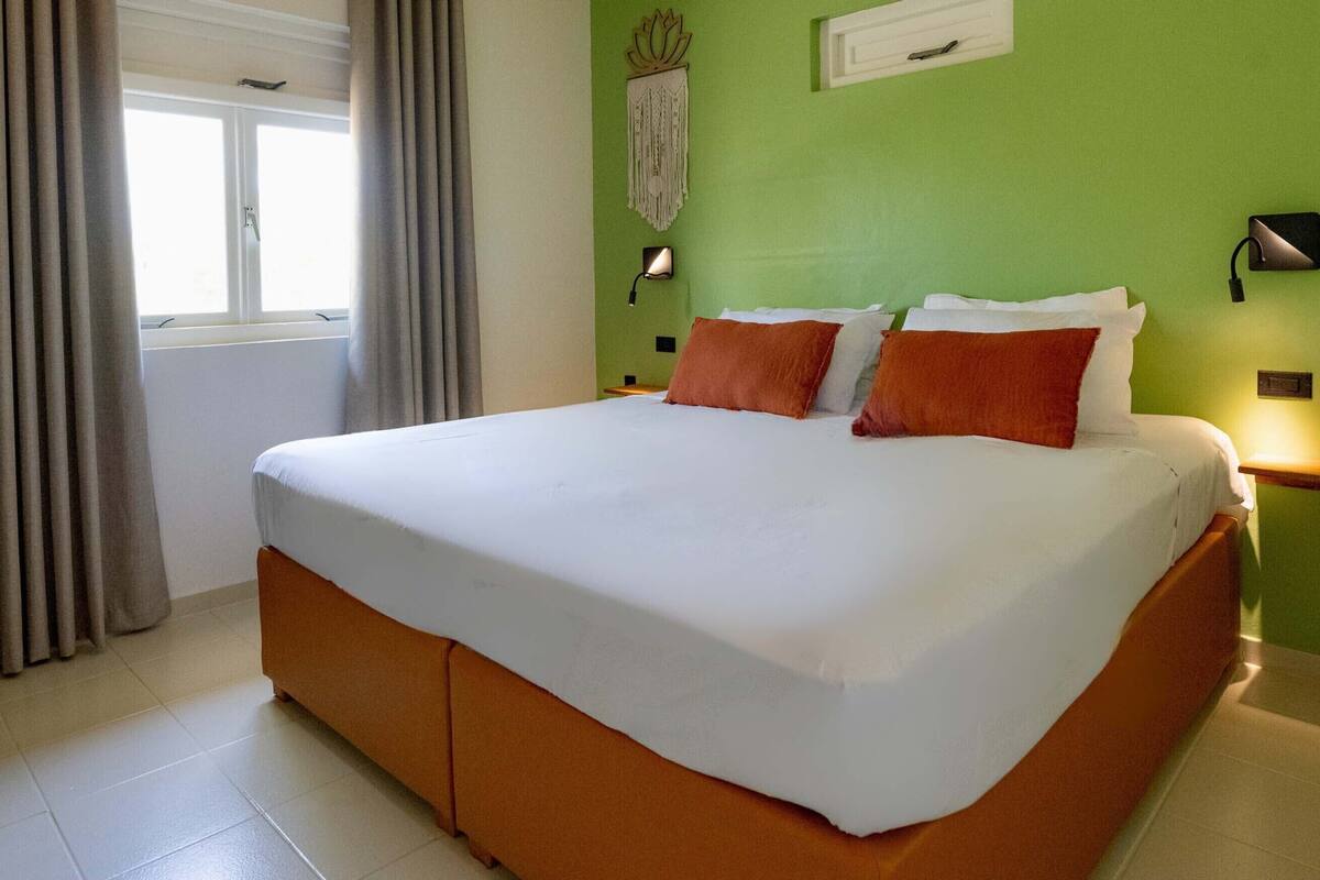 A hotel room with a large bed, grey curtains, a window, a wall-mounted light, and a green wall.
