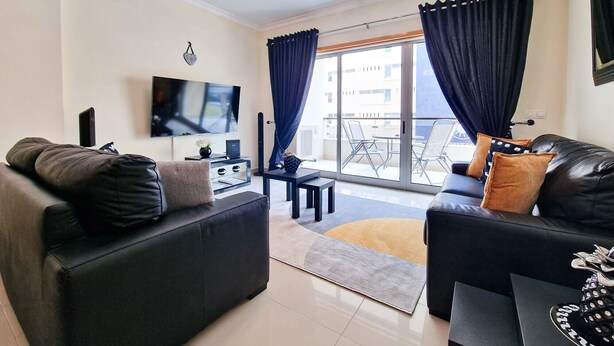 A modern living room with two black leather sofas, a flat-screen TV mounted on the wall, a small side table, and a balcony with a table and chairs.