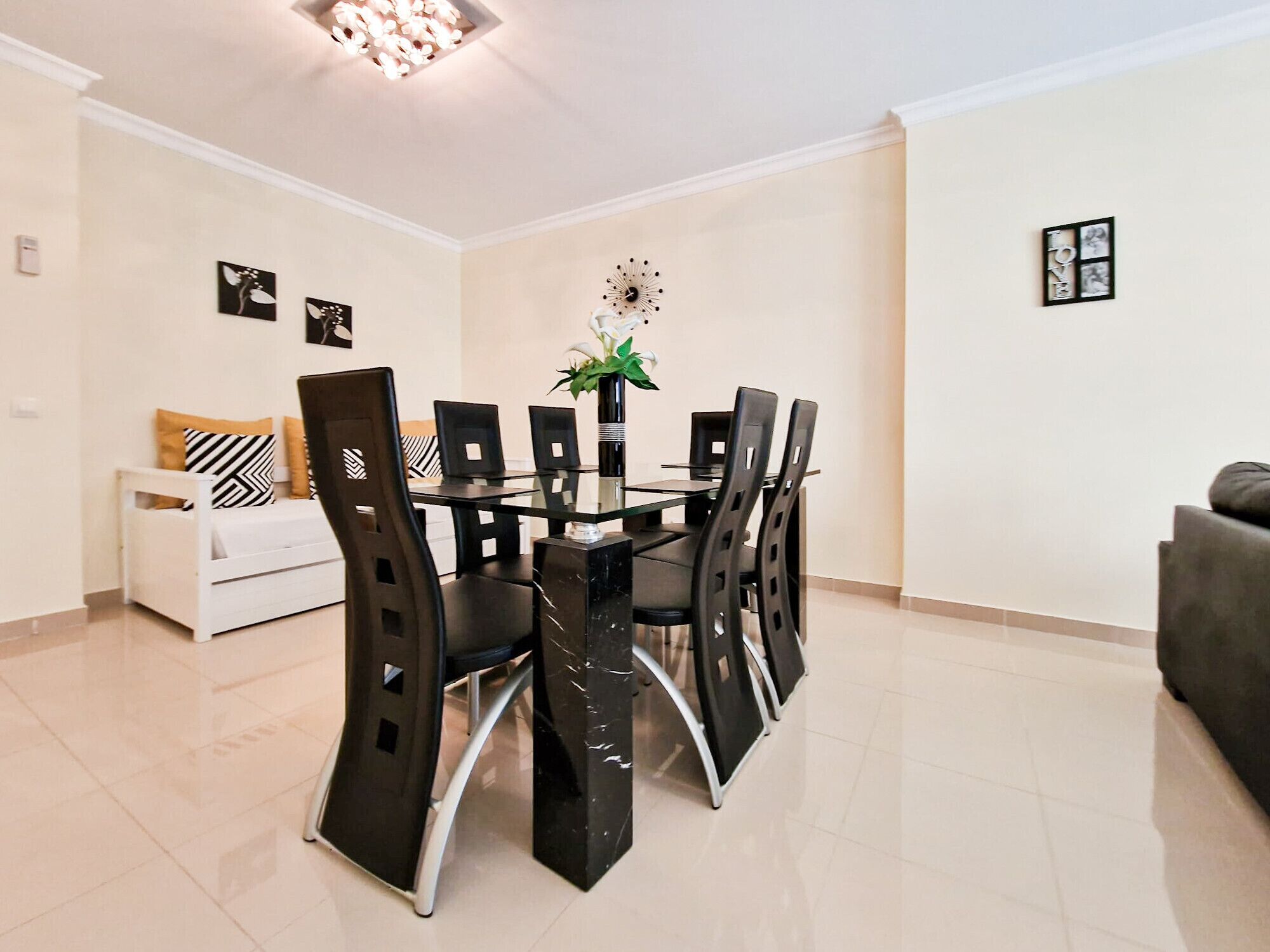 A dining area with a glass table surrounded by chairs, a white sofa, and a small side table with a vase.