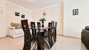 A dining area with a glass table surrounded by chairs, a white sofa, and a small side table with a vase.