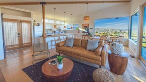 Amplia sala de estar con sofá, mesa de centro y sillones, una cocina al fondo y un ventanal con vistas a la ciudad.