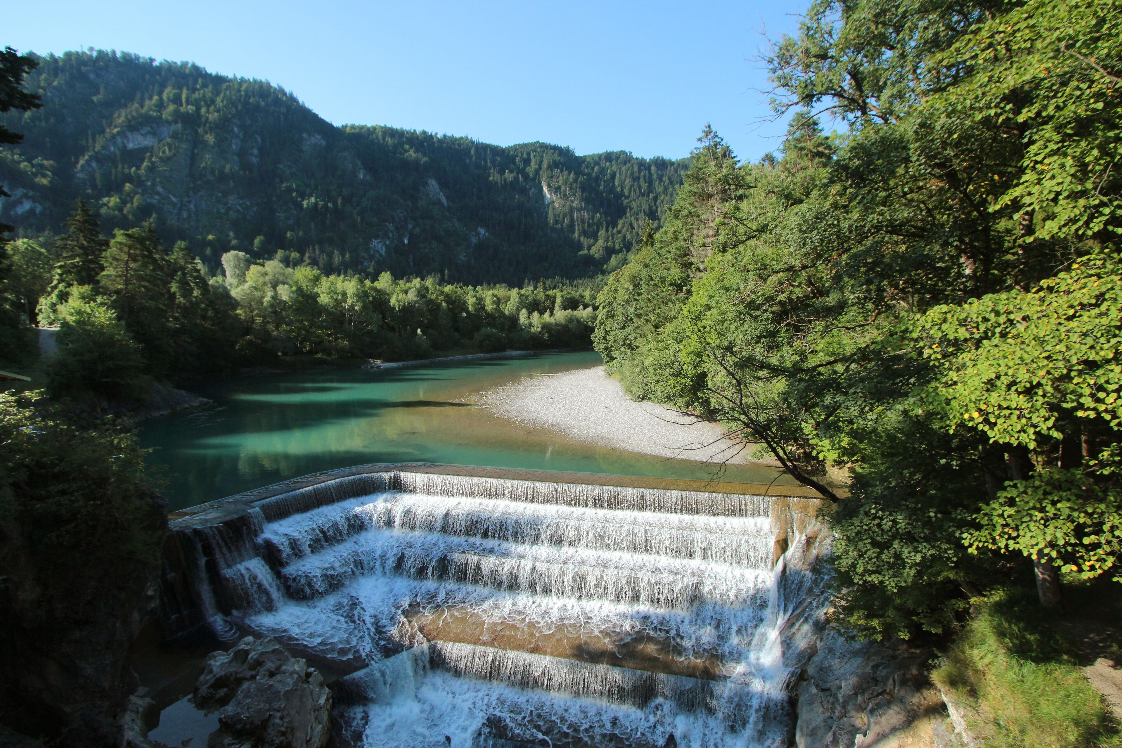 Ein Wasserfall stürzt in einen Fluss, umgeben von dichtem Wald und Hügeln.