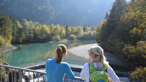 Zwei Personen blicken auf einen Fluss mit einem Wasserfall und bewaldeten Hügeln im Hintergrund.