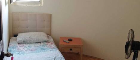 Un dormitorio con cama, mesita de noche, ventilador y ventana.