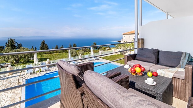 A balcony with a view of the sea, a pool, and a seating area with cushions and a fruit bowl. - Gkountis Deluxe Apartment: Sea View, Shared Pool & Wi-Fi (Lefkadas)