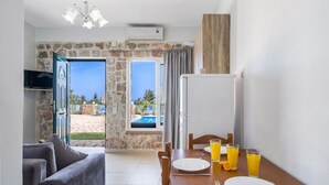 A room with a dining table set for two, a sofa, a TV, and a view of a swimming pool. - Gkountis Stone Apartment 3: Sea View, Shared Pool & Wi-Fi (Lefkadas)